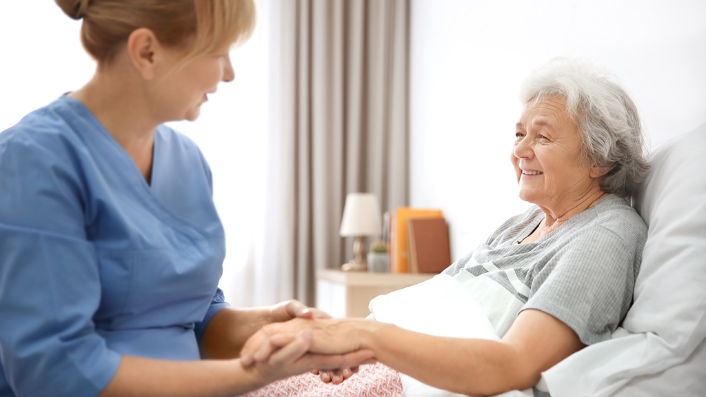 Vergrößerung des Bildes für Krankenschwester am Bett einer älteren Dame in häuslicher Umgebung. Sie hält die Hand der älteren Dame.