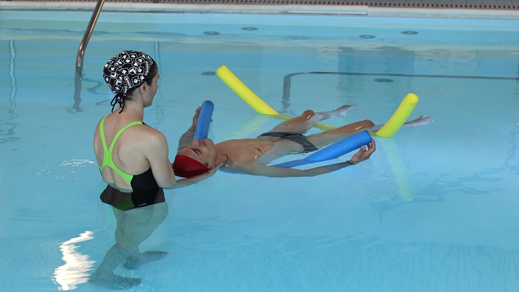 Vergrößerung des Bildes für Zwei Personen im Schwimmbecken; ein Mann liegt auf dem Wasser auf Schwimmnudeln, eine Frau steht im Wasser an seinem Kopfende und stützt seinen Kopf.