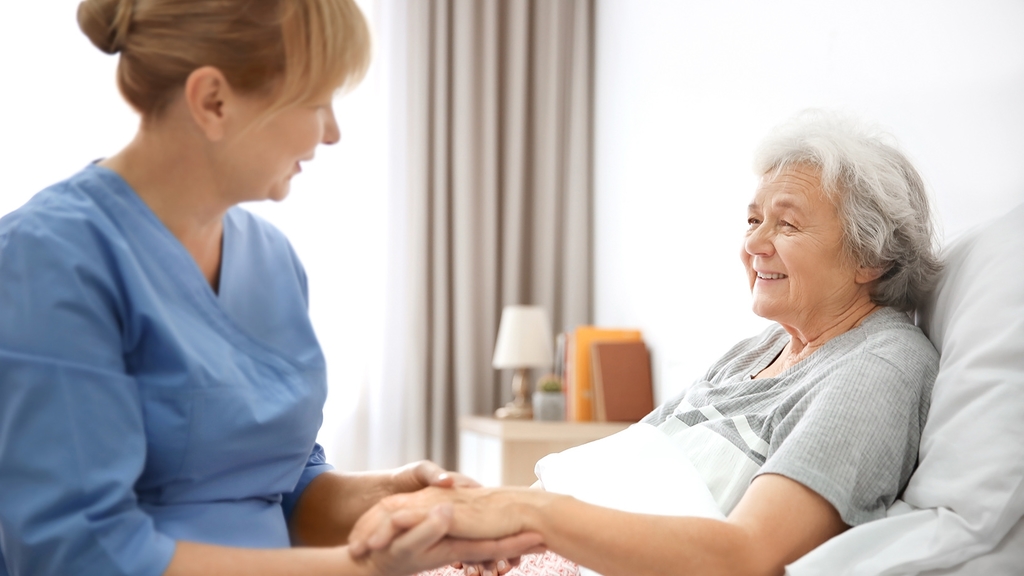 Vergrößerung des Bildes für Pflegeperson sitzt am Bett einer alten Dame und hält ihre Hand.