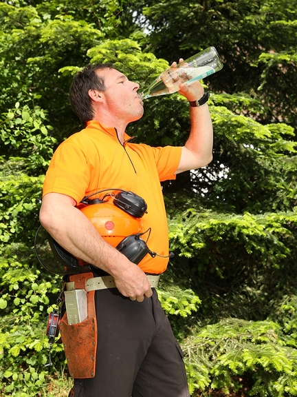 Vergrößerung des Bildes für Ein Mann in Arbeitskleidung und mit Helm unter dem Arm trinkt draußen vor Bäumen aus einer Flasche Wasser..
