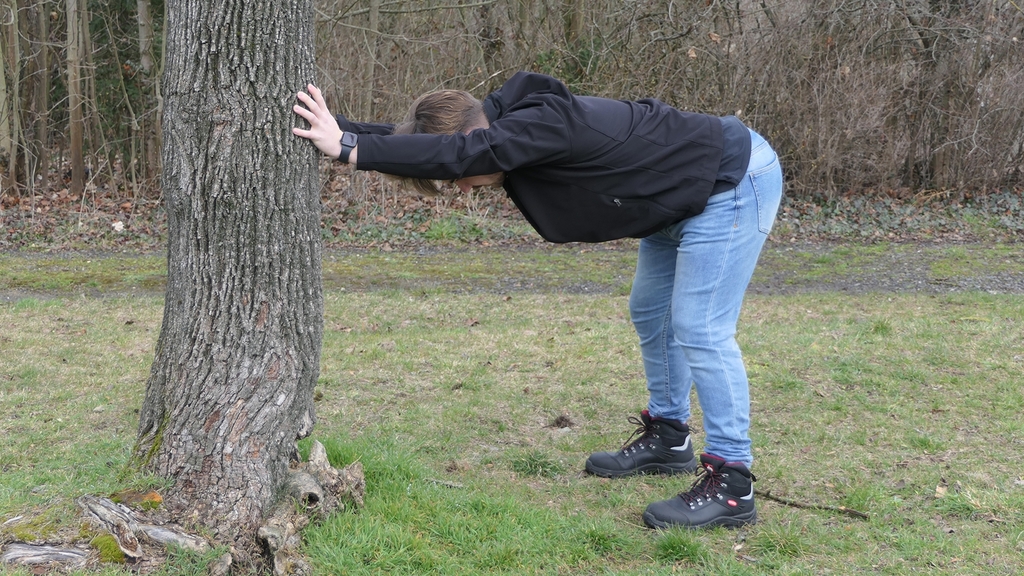Junger Mann macht Bewegungsübung in der Natur an einen Baum abstützend.