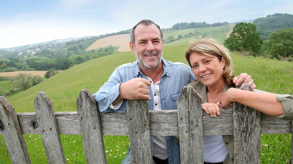 Vergrößerung des Bildes für Ehepaar mittleren Alters steht nebeneinander an einen Zaun gelehnt in ländlicher Umgebung. Im Hintergund sind Wiesen und leichte Hügel zu sehen. .