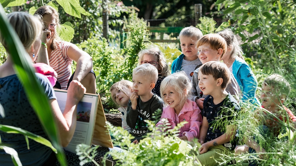 Eine Lehrerin unterrichtet eine Gruppe von Kindern, umgeben von einem Garten, 