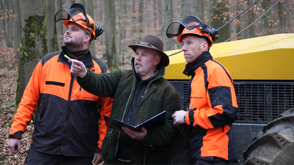 Ein Förster und zwei Beschäftigte im Wald: Abstimmung der Arbeitsschutzerfordernisse. Daneben steht die Forstmaschine.