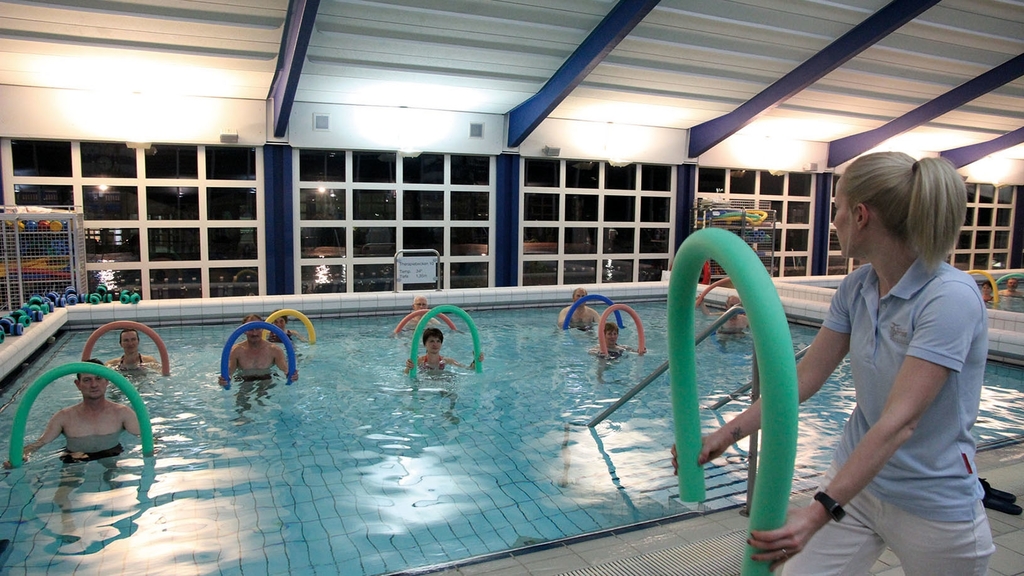Vergrößerung des Bildes für Eine Übungsleiterung steht am Beckenrand eines Schwimmbades mit einer Schwimmnudel. Im Schwimmbecken sind mehrere Personen, die danach angeleitet werden ihre Übungen durchzuführen..