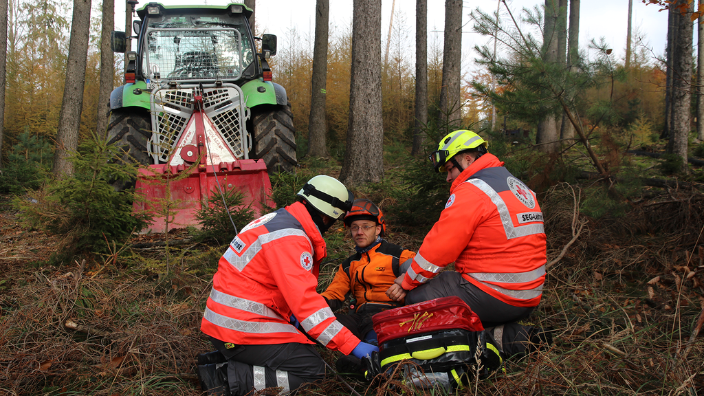 Vergrößerung des Bildes für Ein Waldarbeiter liegt auf dem Waldboden, Zwei Rettungssanitäter knien vor ihm. Im Hintergrund steht ein Traktor..