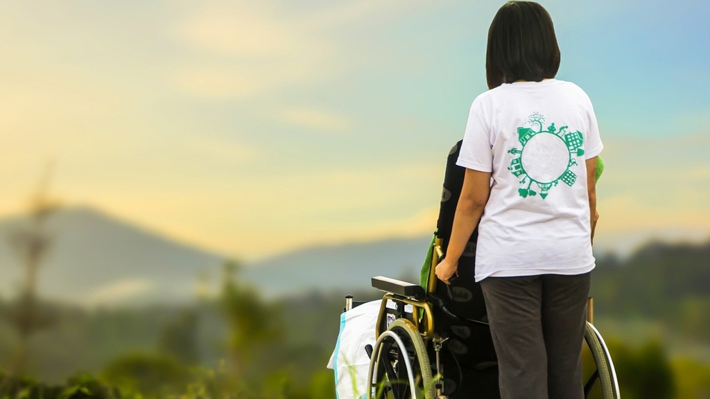 Rückenansicht einer jungen Frau, die - mit einem Patienten im Rollstuhl in der Natur stehend - die Aussicht auf eine Berglandschaft genießt
